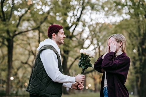 Free Side view of young loving man giving white roses as surprise present to girlfriend covering eyes with hands during date in park Stock Photo