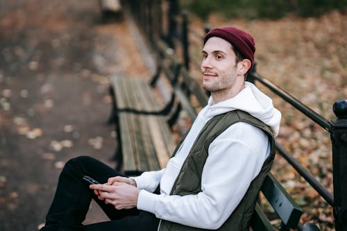 From above of content young male with beard sitting on bench in park and looking away while text messaging on smartphone