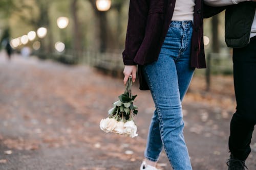 Crop anonymous female in jeans with bouquet of fresh white roses walking in park on blurred background