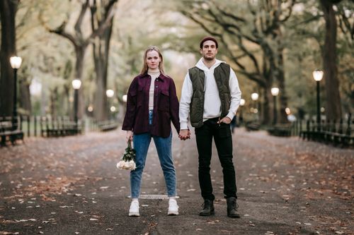 Free Full body of couple with bouquet of white roses in alley covered with leaves near luminous lights and trees Stock Photo