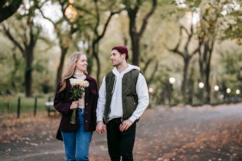 Free Delighted couple with roses walking on alley with trees Stock Photo
