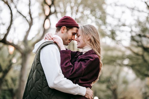 Side view of cheerful couple in casual outfit cuddling and smiling on blurred background of trees in park