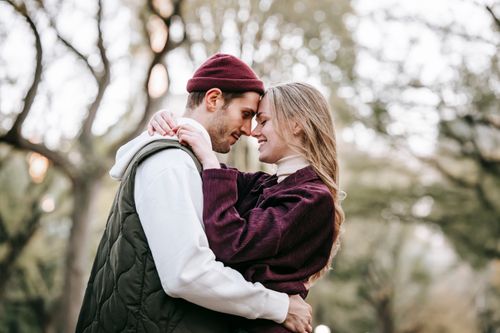 Free Side view of cheerful couple in casual outfit cuddling and smiling on blurred background of trees in park Stock Photo