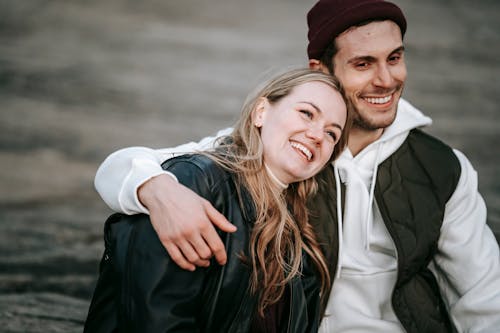 Crop cheerful male in casual apparel embracing young content female beloved while looking away on walkway