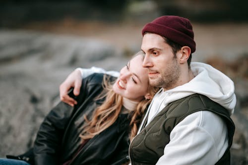 Young wistful male in casual outfit embracing smiling female partner while spending weekend together and looking away