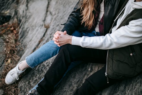 From above of crop unrecognizable couple in casual warm clothes sitting on rocky slope hugging and holding hands