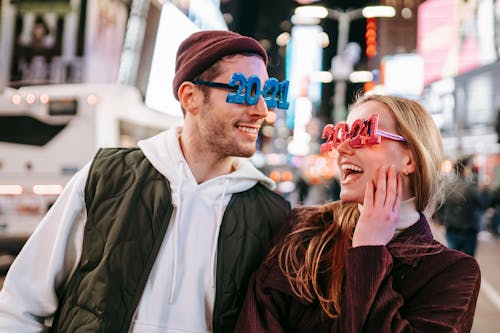 Smiling couple wearing decorated 2021 glasses looking at each other while standing on street with illuminated buildings on blurred background