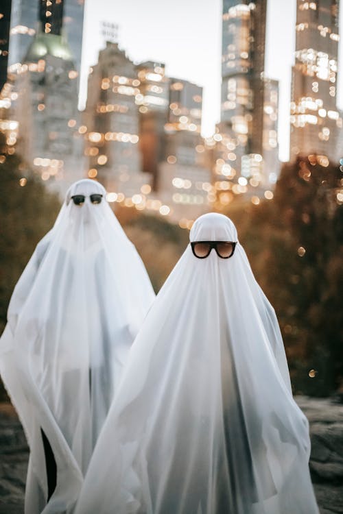 Spooky anonymous couple wearing ghost costumes and sunglasses standing on street with illuminated skyscrapers on blurred background during Halloween holiday