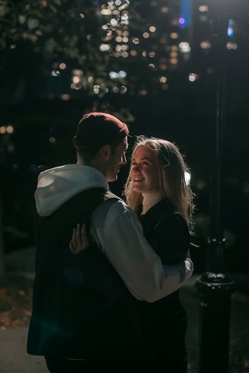 Free Side view of romantic couple cuddling looking at each other while standing on street against blurred trees and buildings at night time Stock Photo