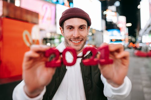 Smiling male wearing hat looking at camera with red decorative glasses with holiday design while standing on street with blurred glowing signboards on modern buildings at night time