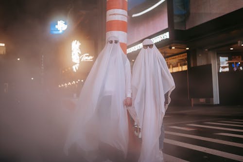 Free Anonymous couple wearing ghost costumes and sunglasses holding hands while standing near steam pipe and building in night city during Halloween Stock Photo