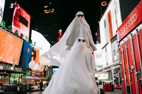 Unrecognizable couple wearing ghost costumes and sunglasses on square with glowing signboards on contemporary buildings at night time on street during Halloween