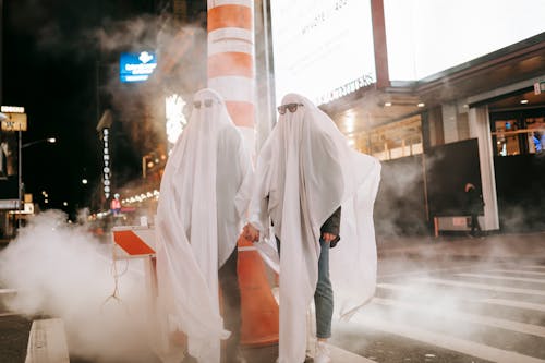 Full body of anonymous couple wearing ghost costumes and sunglasses holding hands while standing on crosswalk near steam pipe at night time