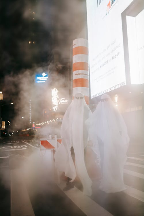 Full body of anonymous couple wearing ghost costumes and sunglasses standing on crosswalk with steam pipe at night city with glowing signboards during Halloween