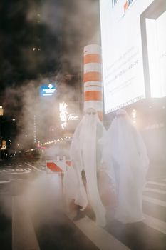 万圣节期间，一对身着幽灵服装、戴着太阳镜的匿名夫妇，站在夜城人行横道上，身旁的蒸汽管冒着气，招牌正发着光