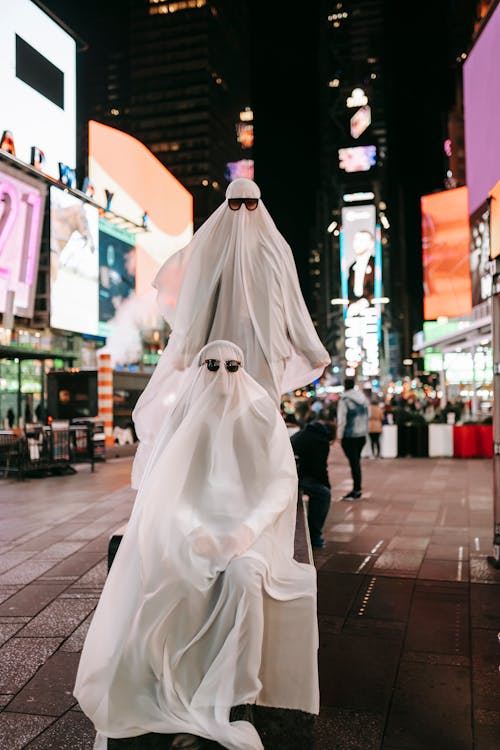 Full body of anonymous couple wearing ghost costumes and sunglasses on stand in night street with glowing signboard on buildings during Halloween