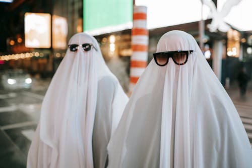 Anonymous couple wearing ghost costumes and sunglasses standing on road near steam pipe and glowing signboards on blurred background on street