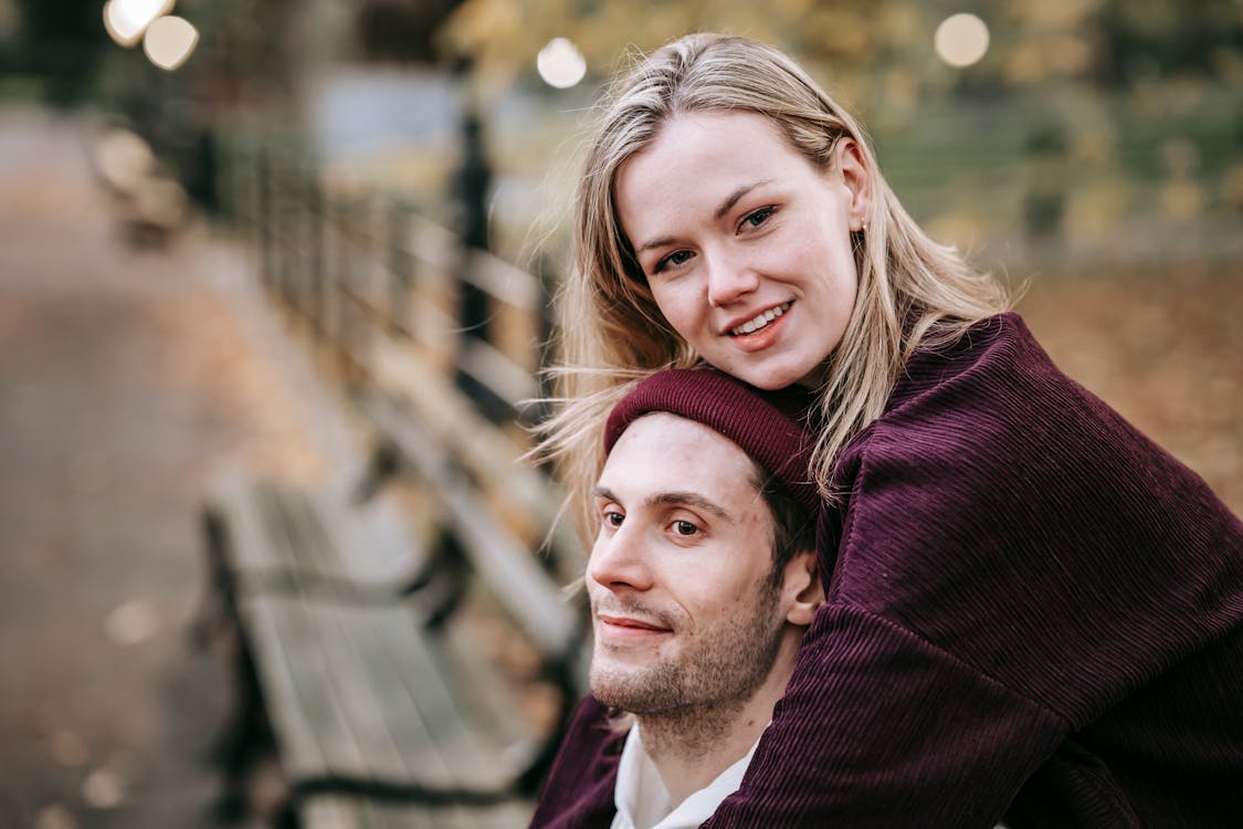 Free Positive couple in casual clothes hugging while sitting on bench in park with autumn trees in daytime Stock Photo