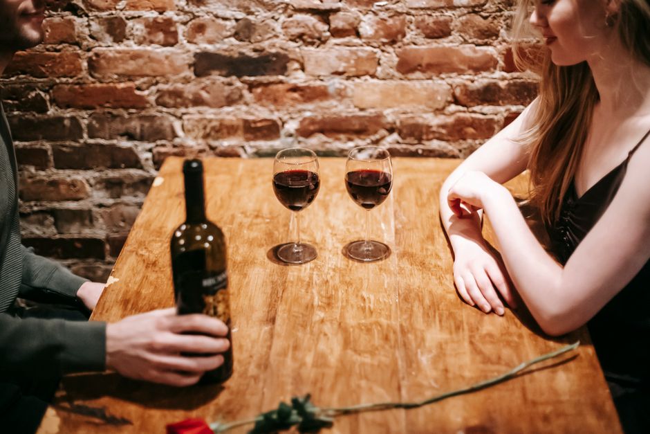 Crop anonymous friends sitting at wooden table with glasses of red wine against brick wall in pub