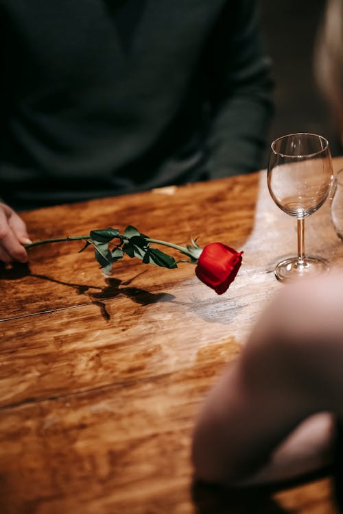 High angle of crop faceless couple sitting in light restaurant at wooden table on romantic date with red rose and wineglasses