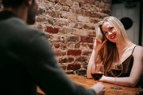 Young couple in elegant outfits sitting in light cafeteria at wooden table near brick wall and having romantic dinner with red wine in glasses and looking at each other happily