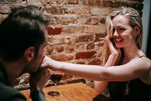 Unrecognizable young man kissing hand of young cheerful blond woman sitting at table in cafe