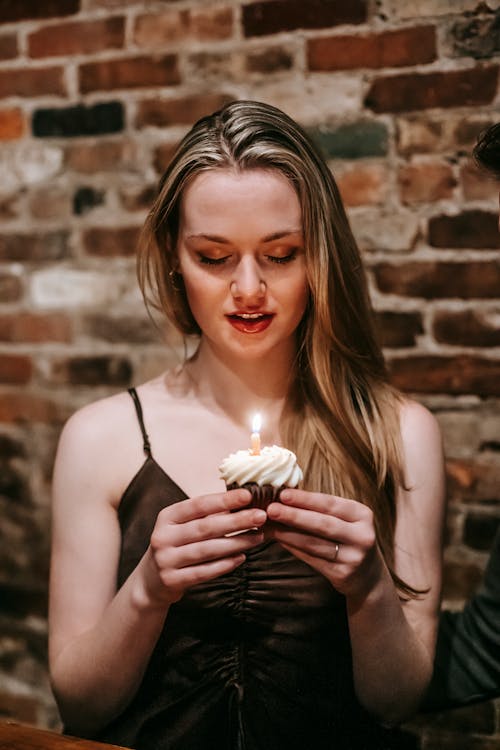 Young blond female with closed eyes holding cupcake with small candle standing near brick wall in room