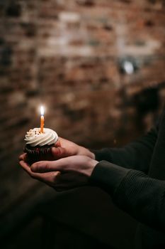 Side view of crop faceless guy in casual outfit standing in light restaurant with small cupcake and flaming candle in hands