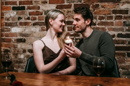 Happy young couple in elegant clothes sitting at table and celebrating birthday with small cupcake and candle near glasses with red wine and brick wall while looking at each other