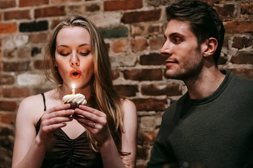 Free Young couple in elegant outfits in restaurant while blowing in candle on small cupcake and celebrating birthday near brick wall Stock Photo
