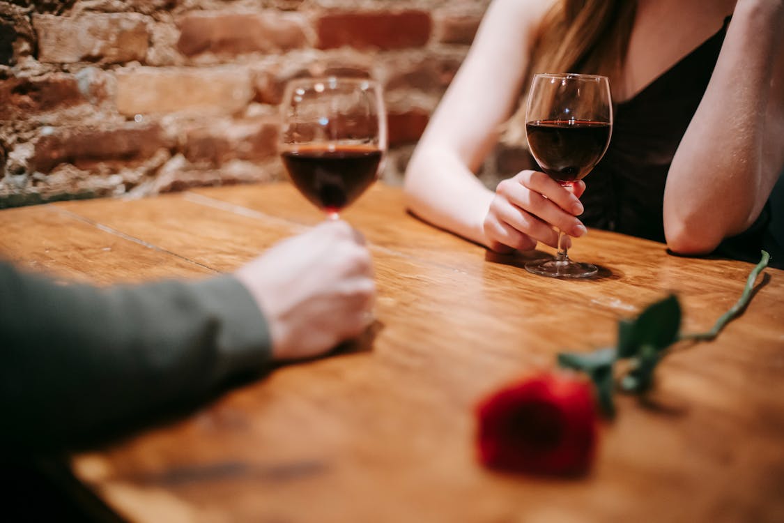 Crop Anonymous Couple In Elegant Outfits Enjoying Romantic Date In Bar At Table With Red Rose And Glasses With Wine Near Brick Wall