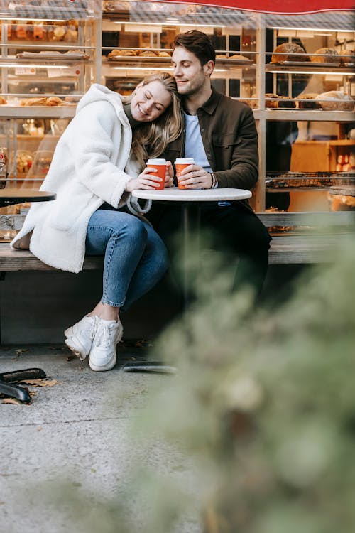 Happy young couple in warm clothes sitting in city near building at round table in street cafe with coffee cups in hands while leaning on shoulder in daylight