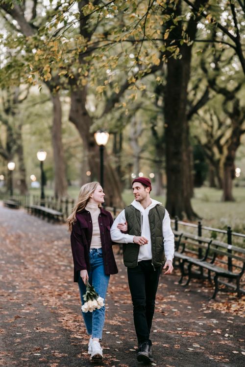 Free Full length of young couple in warm clothes walking arm in arm with bouquet with white flowers in daylight on street near trees in fall and looking at each other Stock Photo