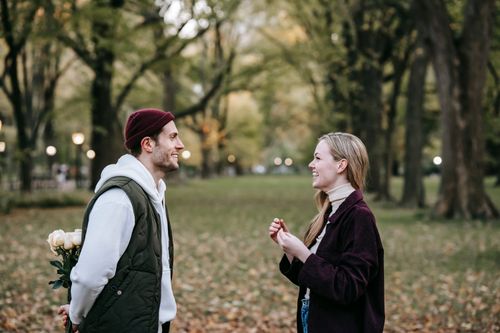 Free Side view of smiling young couple in outerwear standing in park while man preparing to give bouquet of white roses to woman in autumn day while looking at each other Stock Photo