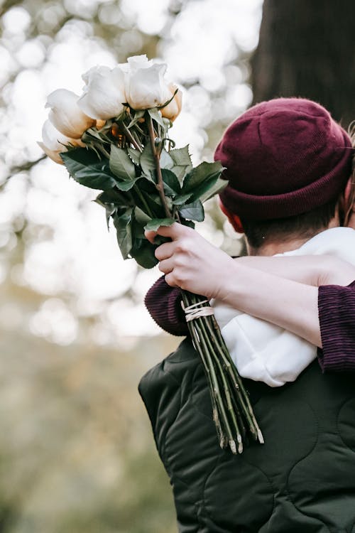 Faceless couple in outerwear standing in park while hugging with bouquet of white roses in hand in daytime