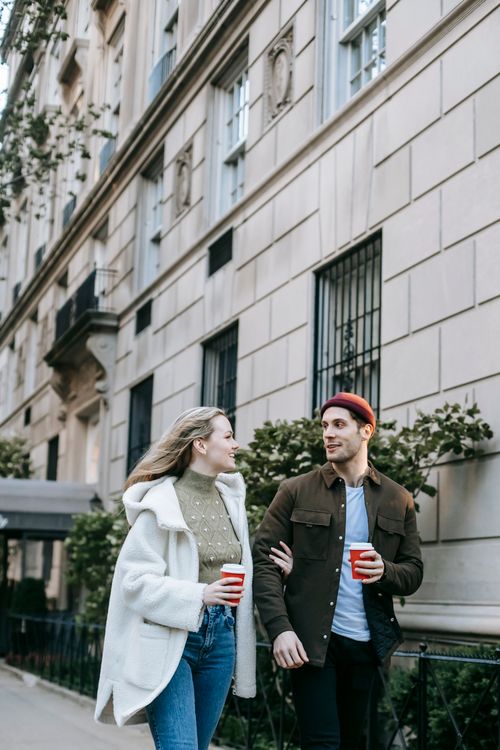 Free Young couple in warm clothes strolling in city street near building on sidewalk with cups of drink in hands and arm in arm and looking at each other in daylight Stock Photo