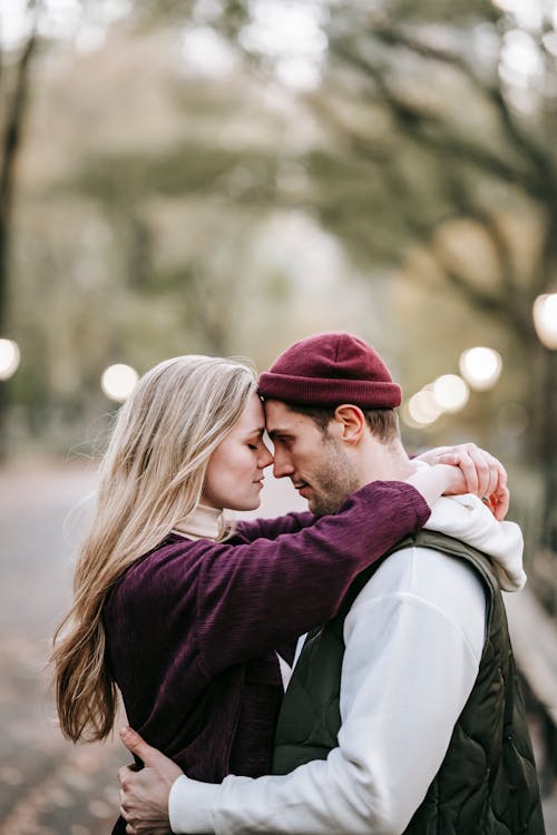 Free Side view of romantic couple in casual outfits standing on blurred background of park and hugging Stock Photo