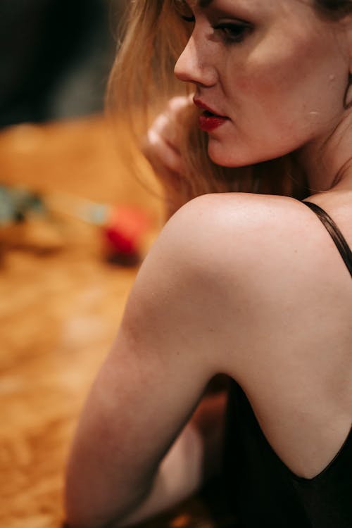 Side view of dreamy female in black outfit sitting at wooden table with red rose on blurred background during romantic date in modern restaurant