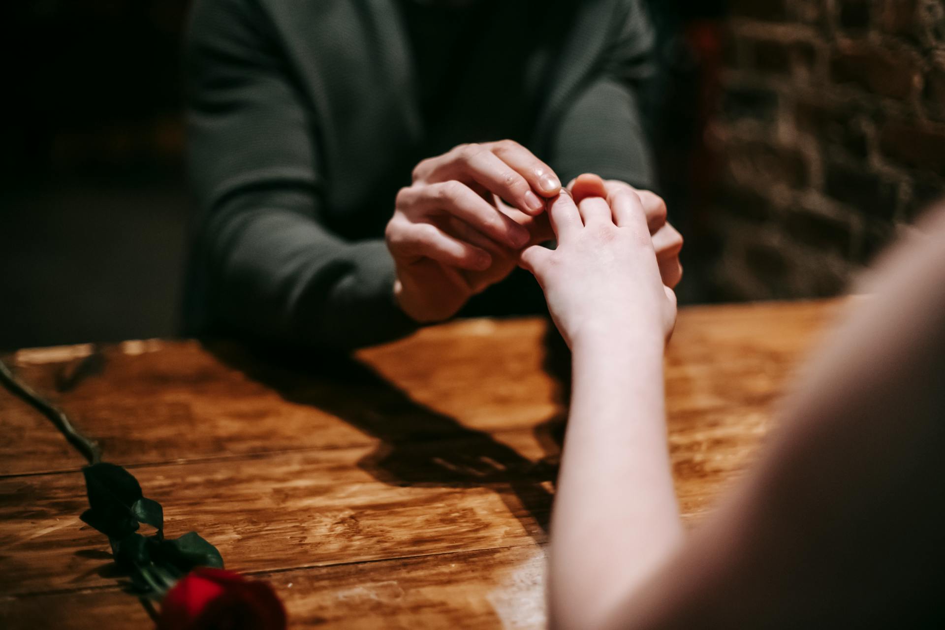 Unrecognizable boyfriend proposing to faceless girlfriend while sitting at wooden table with red rose in modern cafe during romantic date