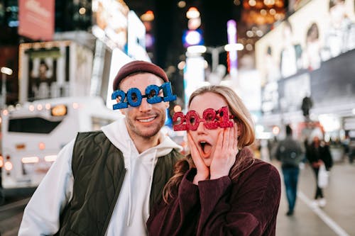 Emotional couple wearing new years 2021 glasses standing near road on square with people and glowing modern buildings on blurred background in downtown at night time