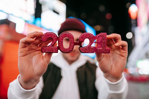 Anonymous male in hat demonstrating red new years 2021 glasses while standing on street with blurred signboards on modern buildings at night time