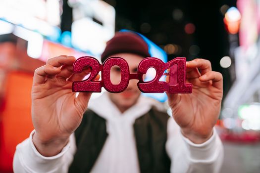 2021年新年夜，一名戴帽子的匿名男子站在现代建筑标牌模糊的街道上，展示红色新年眼镜
