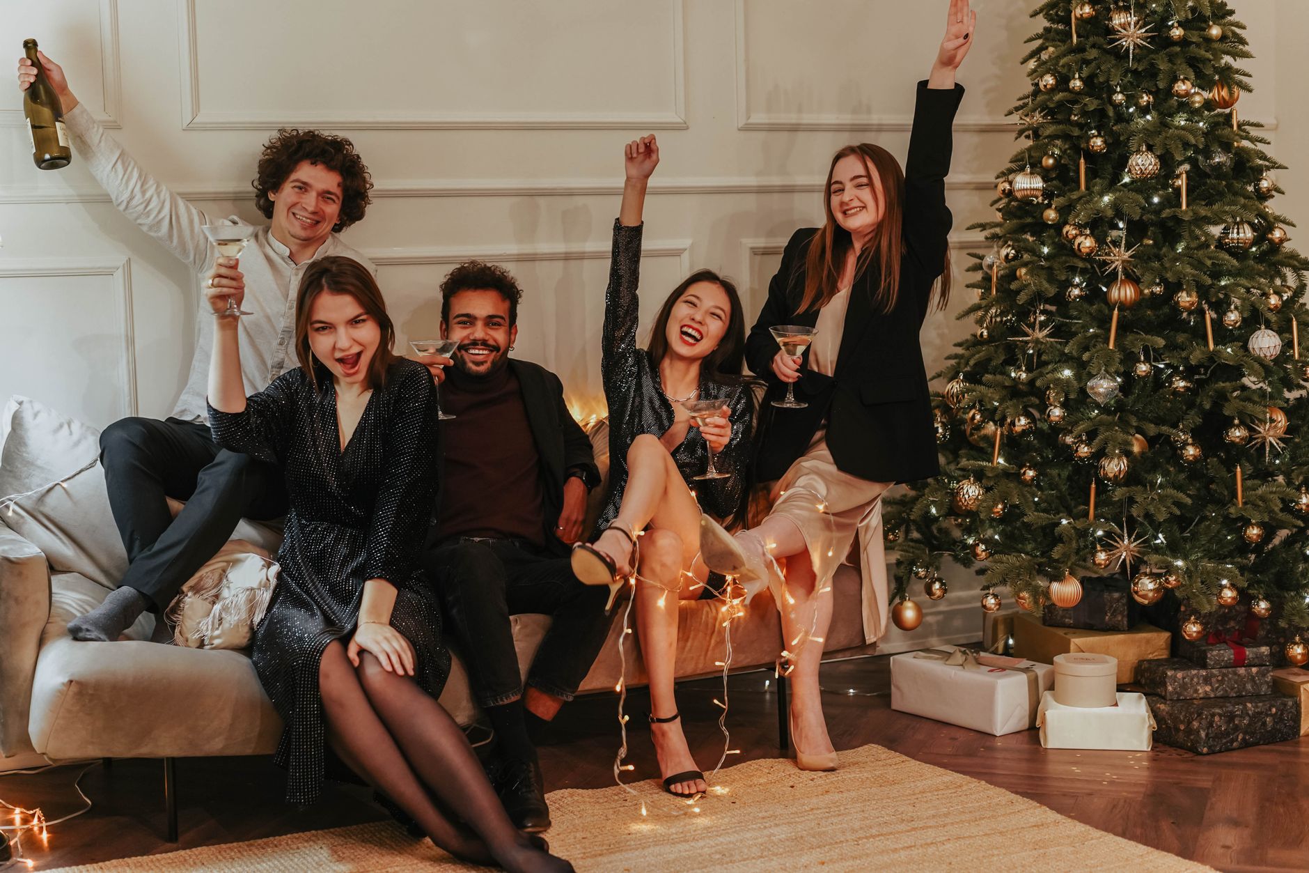 A joyful group of adults celebrating with champagne and laughter indoors by a decorated Christmas tree.