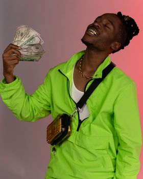 Young man in neon jacket joyfully holds cash, showcasing fashion and wealth in a vibrant studio shoot.