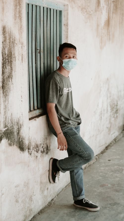 Free Side view of unrecognizable young Asian male millennial with dark hair in casual outfit and medical mask leaning on shabby wall of aged building and looking at camera Stock Photo
