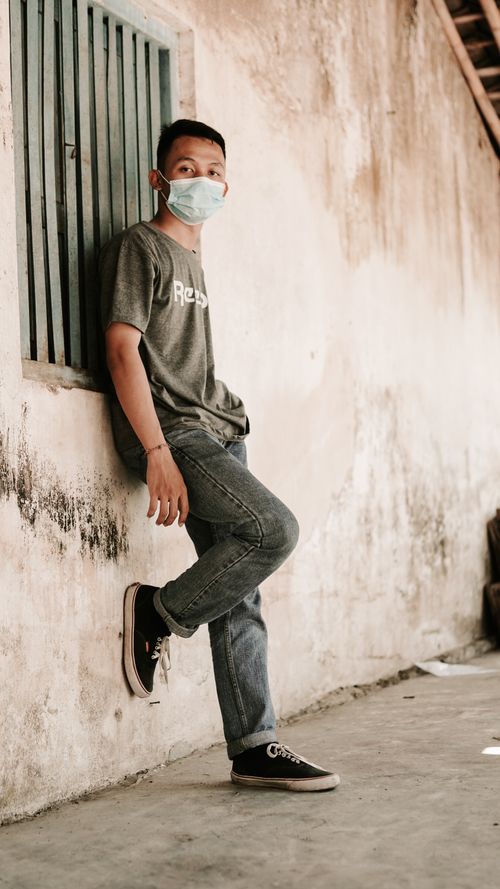 Free Side view full body of Asian teen male with dark hair in casual clothes and protective mask leaning on shabby building wall and looking at camera in suburb Stock Photo