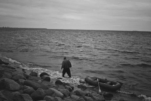 From above black and white of full body anonymous male in black outfit with inflatable boat walking along coast of sea with stones