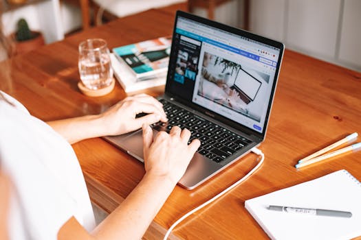 A person working remotely on a laptop at a wooden desk with stationery and a notepad.