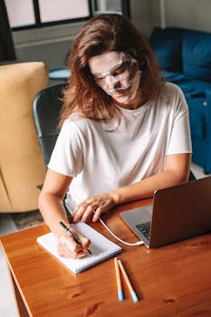 A woman multitasks at home with a face mask, writing notes, and using a laptop.