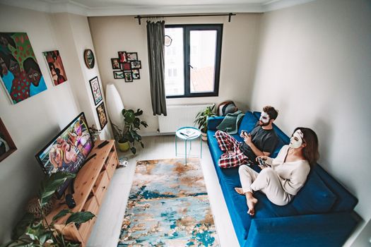 A couple playing video games on a couch with face masks, enjoying leisure time in a cozy living room.
